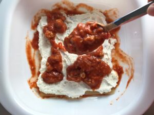 Spoon adding tomato sauce to layer of ricotta in dish for this Easy Italian Sausage Chicken Lasagna Recipe. It's the stuff that dreams are made of. Layers of pasta, creamy ricotta, salty chewy mozzarella, salty and hearty Italian Sausage, and sweet and tangy tomato sauce.