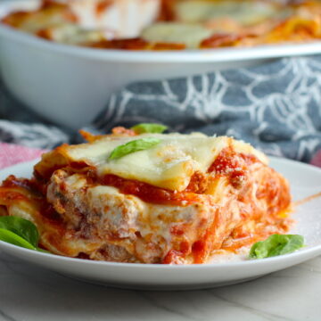 Ground Chicken Lasagna on a plate with fresh basil with casserole dish in background. from the dish. This Italian Sausage-style chicken Lasagne Recipe is the stuff that dreams are made of. Layers of pasta, creamy ricotta, salty chewy mozzarella, salty and hearty Italian Sausage, and sweet and tangy tomato sauce.
