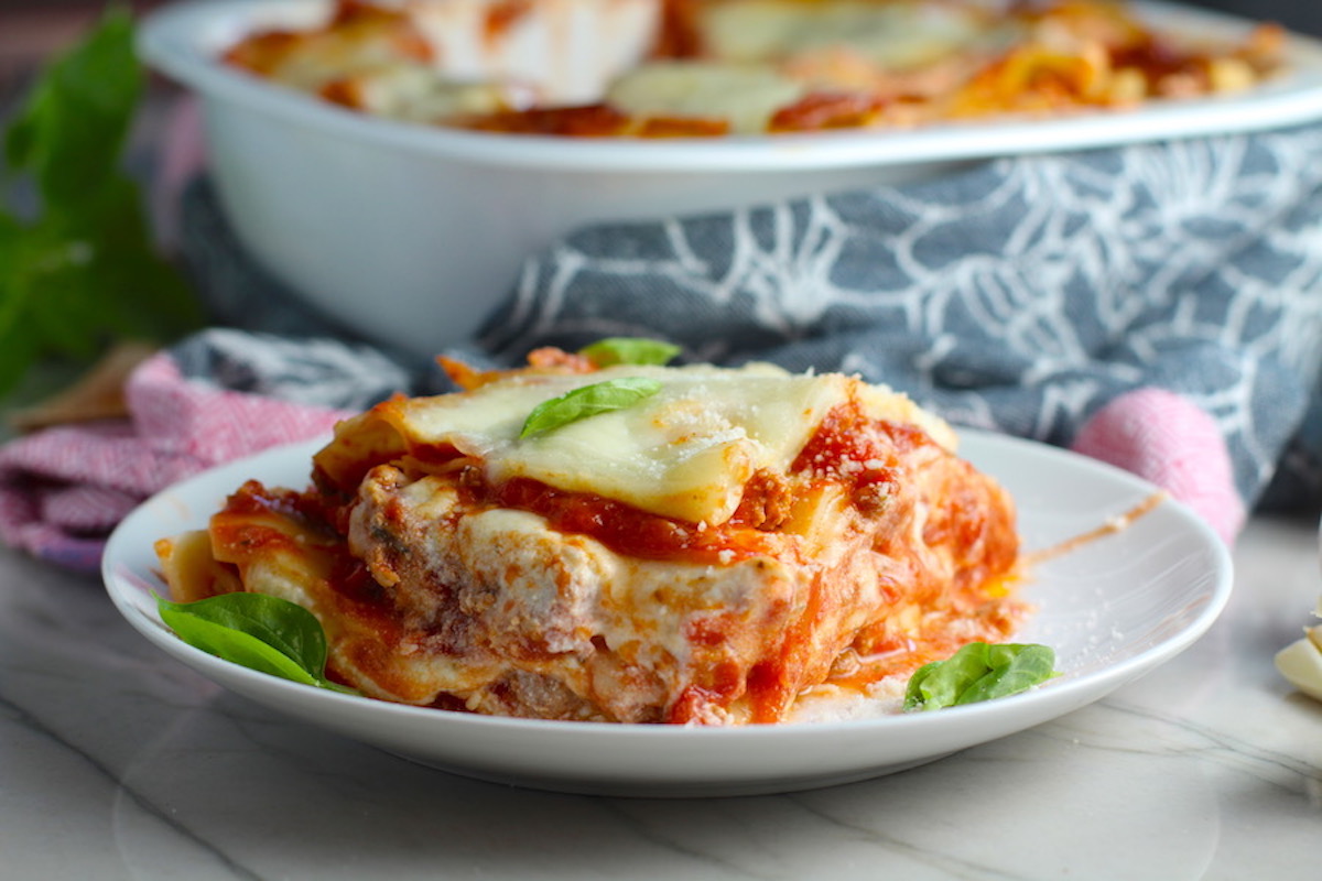 Ground Chicken Layered Lasagna Recipe on a plate with fresh basil with casserole dish in background. from the dish. This Italian Sausage-style chicken Lasagne Recipe is the stuff that dreams are made of. Layers of pasta, creamy ricotta, salty chewy mozzarella, salty and hearty Italian Sausage, and sweet and tangy tomato sauce.