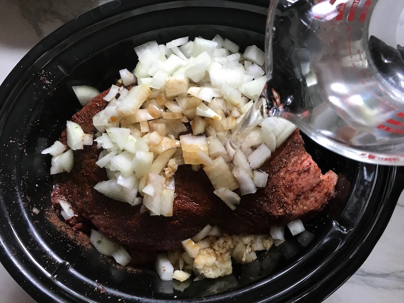 Adding water to onions and raw brisket in Slow Cooker insert for Smoky Pulled Beef Brisket. It's smokey, savory, tender, moist, and full of flavor. In a delicious sauce, it goes great on a sandwich, over polenta, on rice, or potatoes. #slowcooker #crockpot #brisket #beefbrisket #pulledbeef #pulledpork