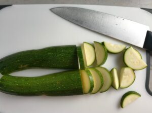 Sliced zucchini on cutting board with knife for Mediterranean SHEET PAN DINNER with chicken sausage, parmesan zucchini. and crispy chickpeas. Serve over quinoa with dollops of seasoned Ricotta. #sheetpandinner #chickpeas