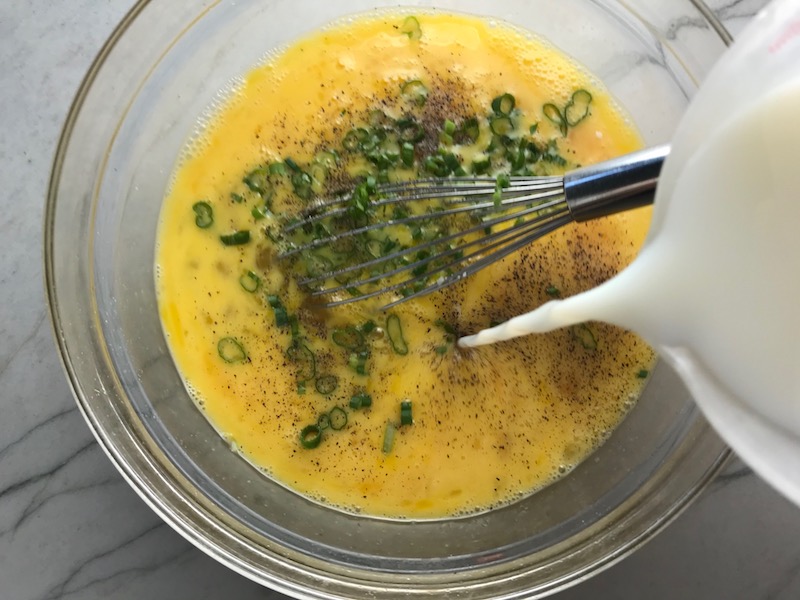 Pouring milk into eggs with scallions in bowl for Slow Cooker Egg Casserole with Chorizo, Potatoes and both cheddar and manchego cheeses. IT COOKS WHILE YOU SLEEP! #eggs #eggcasseroles #eggrecipes #breakfastideas #holidayrecipe