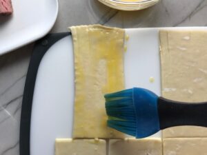Brushing Puff Pastry sheet with egg for Mini Beef Wellington Bites. They are flaky and buttery Puff Pastry is filled with a creamy mushroom and parmesan filling and tender beef filet. #appetizers #partyfood