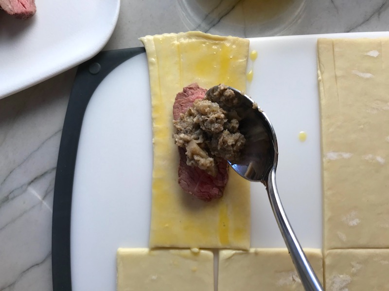 Spoon adding mushroom filling on top of slice of beef on cut Puff Pastry for Mini Beef Wellington Bites. They are flaky and buttery Puff Pastry is filled with a creamy mushroom and parmesan filling and tender beef filet. #appetizers #partyfood