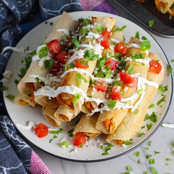 Baked Spicy Chicken Taquitos stacked on plate with toppings. Corn tortillas are filled with smoky taco seasoned shredded chicken, and creamy cheddar cheese.