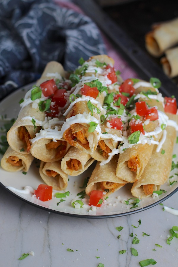 Baked Chicken Taquitos stacked on plate with toppings. Corn tortillas are filled with smoky taco seasoned shredded chicken, and creamy cheddar cheese. #dinnerideas #familydinner #chickenrecipes #chickendinner #tacos #taquitos