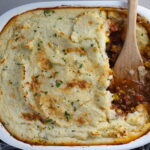 Spatula in Cottage Pie in casserole dish. This recipe has Spanish Chorizo and ground beef cooked in a rich and savory gravy with veggies and herbs.  Creamy mashed potatoes sit on top with manchego, parmesan, and garlic. #dinnerideas #cottagepie #shepherdspie
