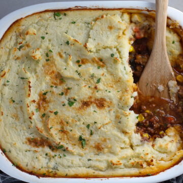 Spatula in Cottage Pie in casserole dish. This recipe has Spanish Chorizo and ground beef cooked in a rich and savory gravy with veggies and herbs. Creamy mashed potatoes sit on top with manchego, parmesan, and garlic. #dinnerideas #cottagepie #shepherdspie