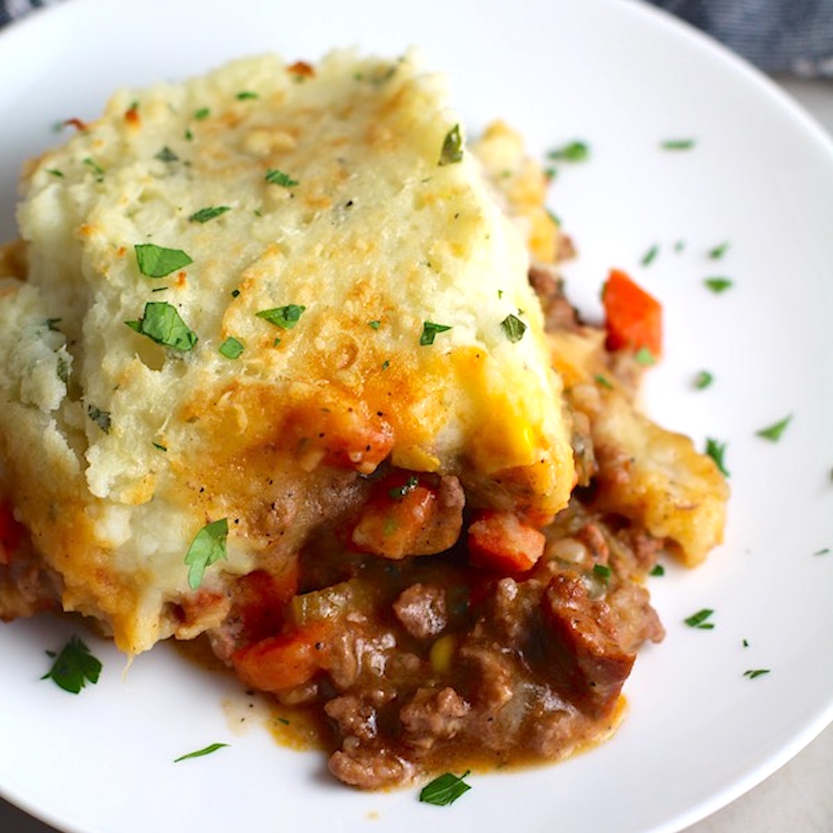 Piece of Sausage Shepherds Pie on plate with dish in back. This recipe has Spanish Chorizo and ground beef cooked in a rich and savory gravy with veggies and herbs. Creamy mashed potatoes sit on top with manchego, parmesan, and garlic.