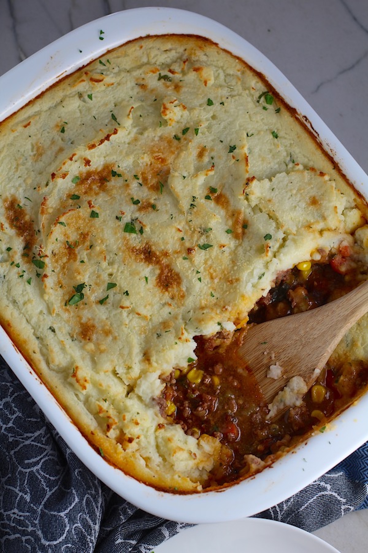 Spatula in Sausage Shepherds Pie in casserole dish. This recipe has Spanish Chorizo and ground beef cooked in a rich and savory gravy with veggies and herbs. Creamy mashed potatoes sit on top with manchego, parmesan, and garlic.