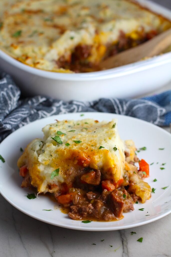 Piece of Sausage Shepherds Pie on plate with dish in back. This recipe has Spanish Chorizo and ground beef cooked in a rich and savory gravy with veggies and herbs.  Creamy mashed potatoes sit on top with manchego, parmesan, and garlic.