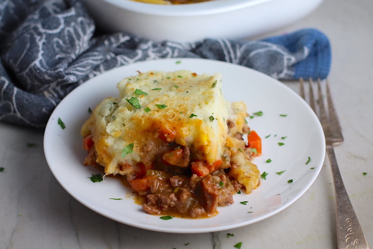 Piece of Sausage Shepherds Pie on plate with dish in back. This recipe has Spanish Chorizo and ground beef cooked in a rich and savory gravy with veggies and herbs. Creamy mashed potatoes sit on top with manchego, parmesan, and garlic.