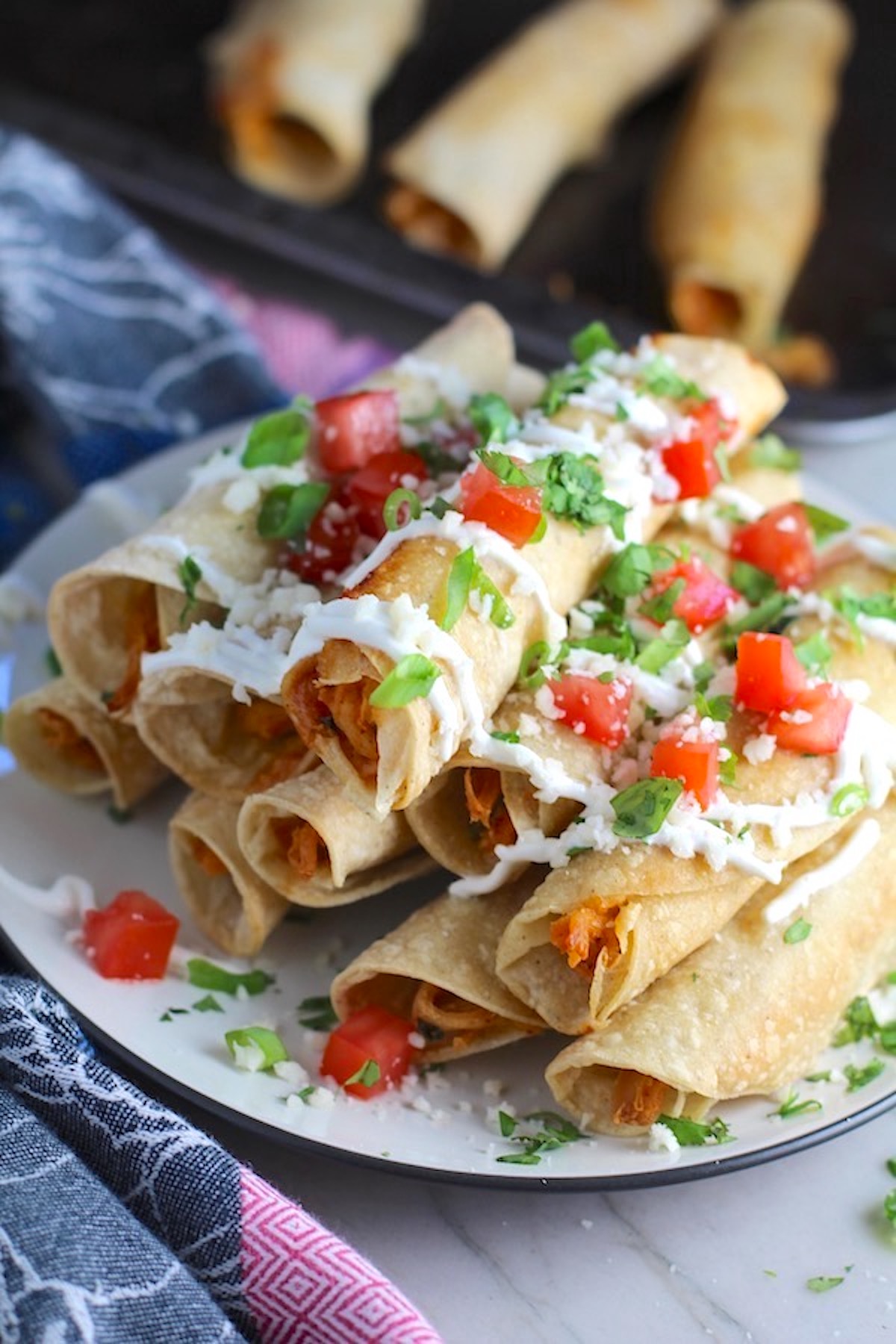 Baked Spicy Chicken Taquitos stacked on plate with toppings. Corn tortillas are filled with smoky taco seasoned shredded chicken, and creamy cheddar cheese.