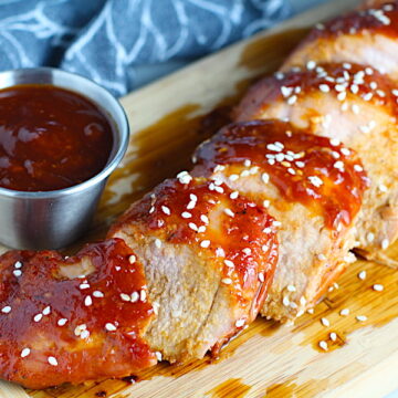 Chinese BBQ Pork Tenderloin sliced on board with sesame seeds and cup of sauce. It's sweet, tangy, salty, nutty, and all together lick-your-fingers delicious! It's also easy to make! Marinade ahead and then roast in the oven.