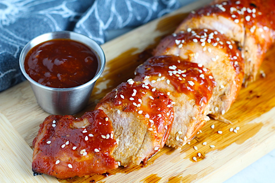 Chinese BBQ Pork Tenderloin sliced on board with sesame seeds and cup of sauce. It's sweet, tangy, salty, nutty, and all together lick-your-fingers delicious! It's also easy to make! Marinade ahead and then roast in the oven.