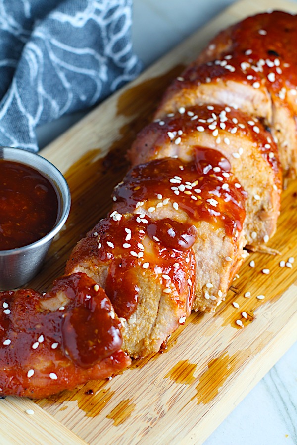 Chinese BBQ Pork Tenderloin sliced on board with sesame seeds and cup of sauce.  It's sweet, tangy, salty, nutty, and all together lick-your-fingers delicious!  It's also easy to make!  Marinade ahead and then roast in the oven. 
