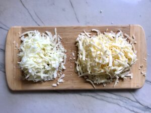 Shredded Manchego and Cheddar on cutting board for Bacon Mac and Cheese. It has a creamy and cheesy sauce with a touch of smokiness from the bacon, coats each piece of pasta. On top, crispy crunchy, indulgent bacon fat cracker crumbs!