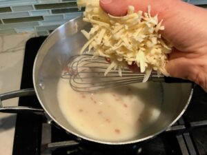 Adding shredded cheese to sauce for Bacon Mac and Cheese. It has a creamy and cheesy sauce with a touch of smokiness from the bacon, coats each piece of pasta. On top, crispy crunchy, indulgent bacon fat cracker crumbs!