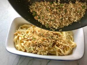 Pouring cracker crumbs over pasta for Bacon Mac and Cheese. It has a creamy and cheesy sauce with a touch of smokiness from the bacon, coats each piece of pasta. On top, crispy crunchy, indulgent bacon fat cracker crumbs!