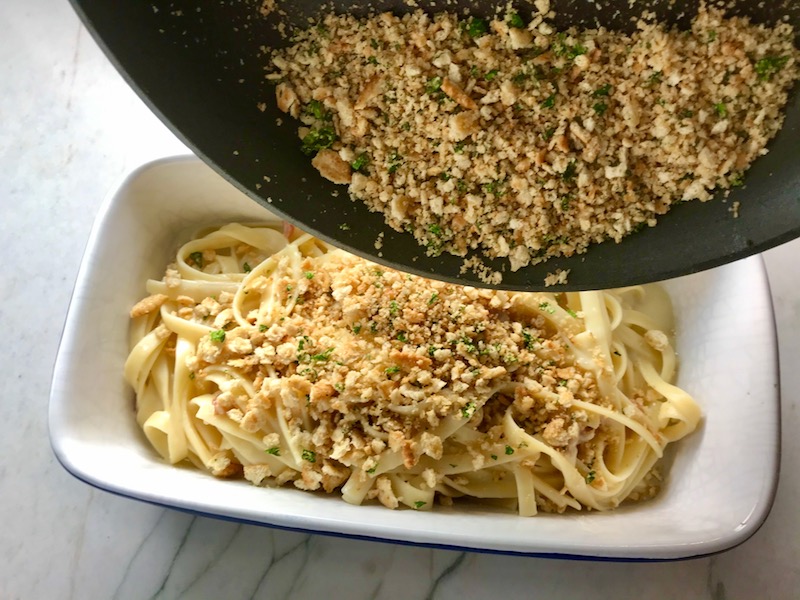 Pouring cracker crumbs over pasta for Bacon Mac and Cheese. It has a creamy and cheesy sauce with a touch of smokiness from the bacon, coats each piece of pasta. On top, crispy crunchy, indulgent bacon fat cracker crumbs!
