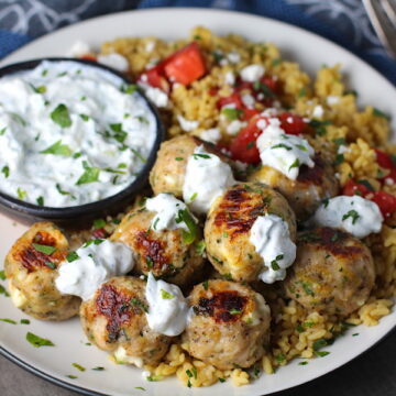 Ground Chicken Greek Meatballs on a plate with Tzatziki sauce over rice. Baked meatballs in back. These meatballs have garlic, feta, parsley, and oregano.
