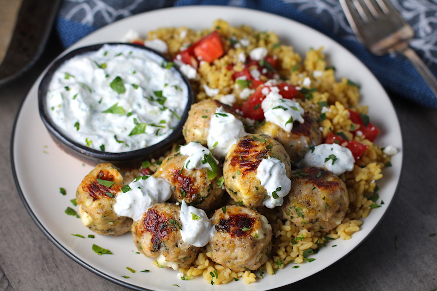 Ground Chicken Greek Meatballs on a plate with Tzatziki sauce over rice. Baked meatballs in back. These meatballs have garlic, feta, parsley, and oregano.