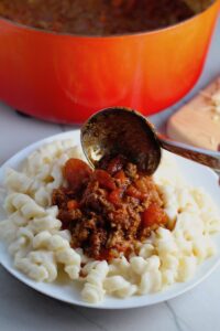 Ladling Chili over Mac and Cheese on a plate. This Recipe has creamy and cheesy pasta topped with smokey, tangy, savory beef chili! #macandcheese #chilirecipes