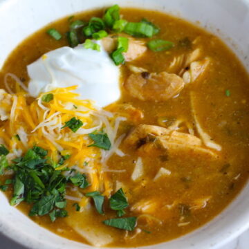Creamy Chicken Green Enchilada Soup in a bowl topped with cheese, sour cream, and cilantro! It's creamy, hearty, slightly spicy with green chiles and smokey seasonings that bring so much depth.  Corn tortilla strips act like the noodles in this soup!