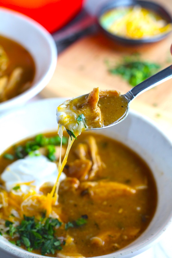 Spoon with Creamy Chicken Enchilada Soup in a bowl topped with cheese, sour cream, and cilantro! It's creamy, hearty, slightly spicy with green chiles and smokey seasonings that bring so much depth. Corn tortilla strips act like the noodles in this soup!