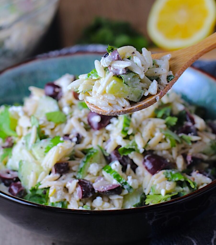 Spoon with scoop of Greek Lemon Orzo Salad with Kalamata Olives, Cucumbers, and Feta Cheese in a bowl with lemon on side. It's mixed with a creamy lemon dressing that's bright and light!