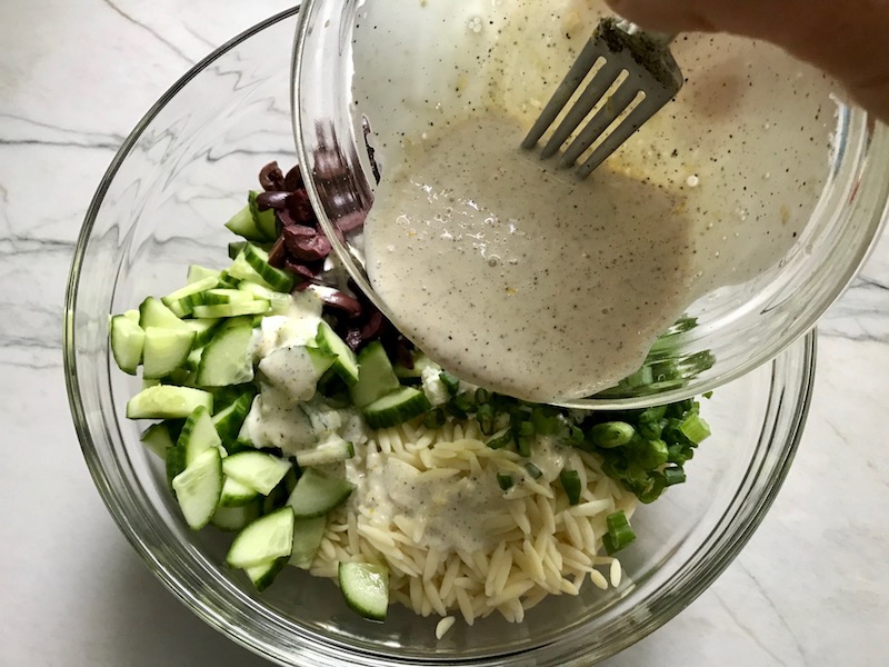 Pouring dressing over Greek Orzo Salad with Kalamata Olives, Cucumbers, and Feta Cheese. It's mixed with a creamy lemon dressing that's bright and light!