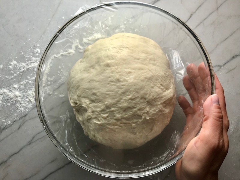Homemade pizza dough in a clear bowl covered with plastic wrap and hand on side for a BBQ Chicken Pizza Recipe