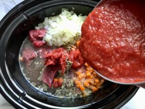 Pouring pureed tomatoes over raw beef, diced onion, carrots, and other ingredients in pot for Shredded Beef Ragu Recipe with Pappardelle. It's so easy to make and has a thick meaty texture and a super rich delicious flavor.
