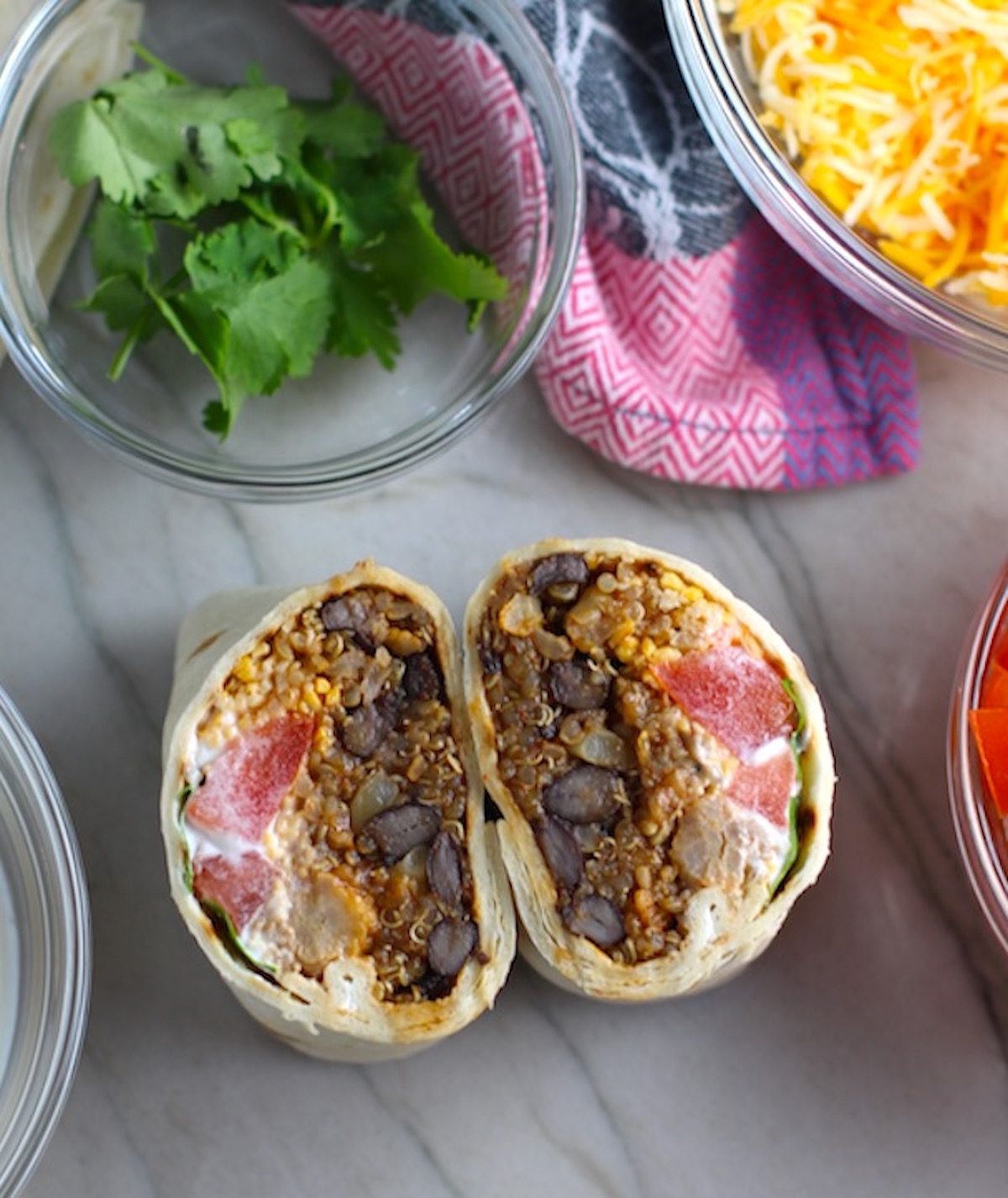 Black Bean and Quinoa Burritos cut in half and standing face up on counter. Packed with protein and fiber, but also cheese, tomato, cilantro, and sour cream! The vegetarian 'meat' is taco-seasoned quinoa, black beans, and cauliflower.