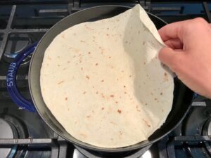 Tortilla in pan for Quinoa Black Bean Burritos. Packed with protein and fiber, but also cheese, tomato, cilantro, and sour cream! The vegetarian 'meat' is taco-seasoned quinoa, black beans, and cauliflower.
