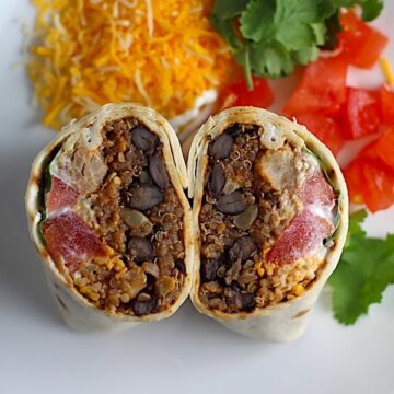 Quinoa Black Bean Burritos cut in half and standing face up on plate. Packed with protein and fiber, but also cheese, tomato, cilantro, and sour cream! The vegetarian 'meat' is taco-seasoned quinoa, black beans, and cauliflower.