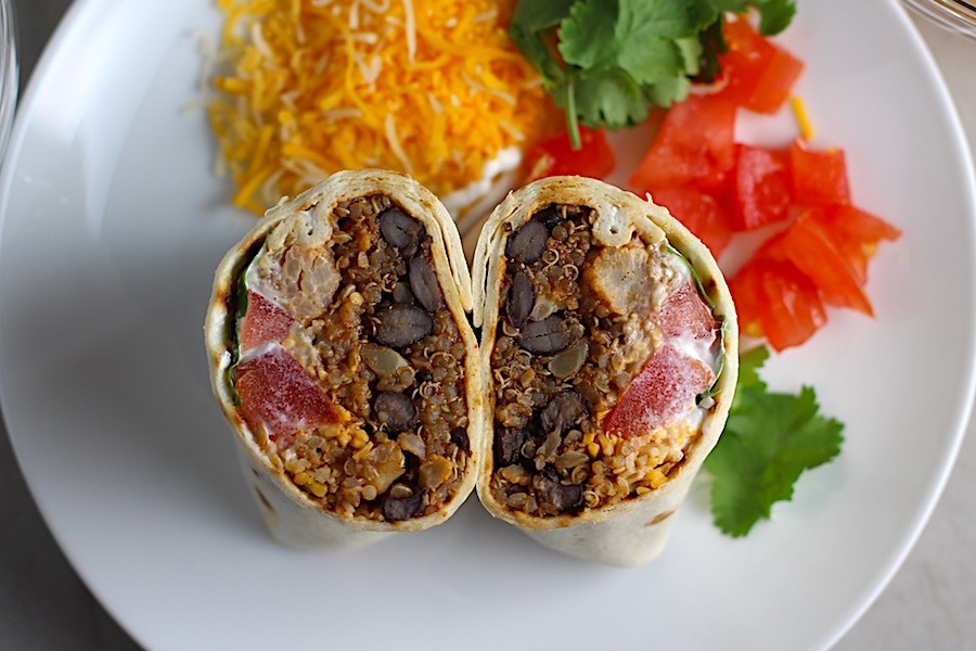 Quinoa Black Bean Burritos cut in half and standing face up on plate. Packed with protein and fiber, but also cheese, tomato, cilantro, and sour cream! The vegetarian 'meat' is taco-seasoned quinoa, black beans, and cauliflower.