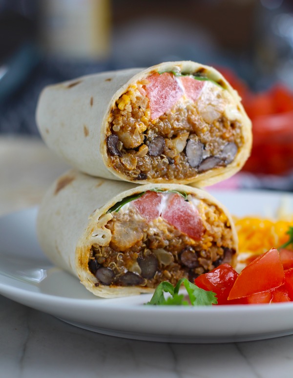 Quinoa Black Bean Burritos cut in half and stacked on plate. Packed with protein and fiber, but also cheese, tomato, cilantro, and sour cream! The vegetarian 'meat' is taco-seasoned quinoa, black beans, and cauliflower.