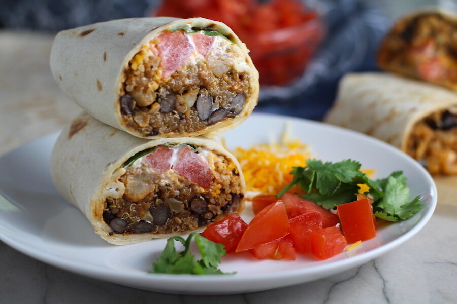 Burritos with Black Beans and Quinoa recipe cut in half and stacked on plate. Packed with protein and fiber, but also cheese, tomato, cilantro, and sour cream! The vegetarian 'meat' is taco-seasoned quinoa, black beans, and cauliflower.