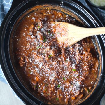 Crockpot Shredded Beef Ragu Recipe in slow cooker with wood spoon and parmesan on the table. It's so easy to make and has a thick meaty texture and a super rich delicious flavor.