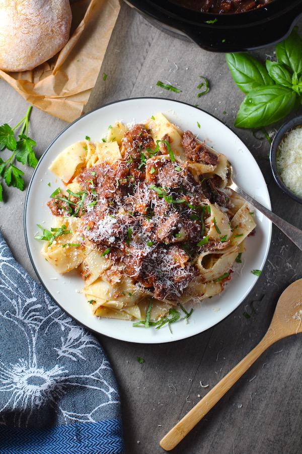 Crockpot Shredded Beef Ragu Recipe with Pappardelle on plate with parsley, basil, parmesan, and bread on the table. It's so easy to make and has a thick meaty texture and a super rich delicious flavor.