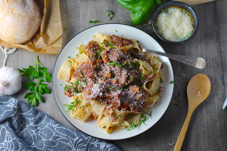 Shredded Beef Ragu Recipe with Pappardelle with a fork on plate with parsley, basil, parmesan, and bread on the table. It's so easy to make and has a thick meaty texture and a super rich delicious flavor.