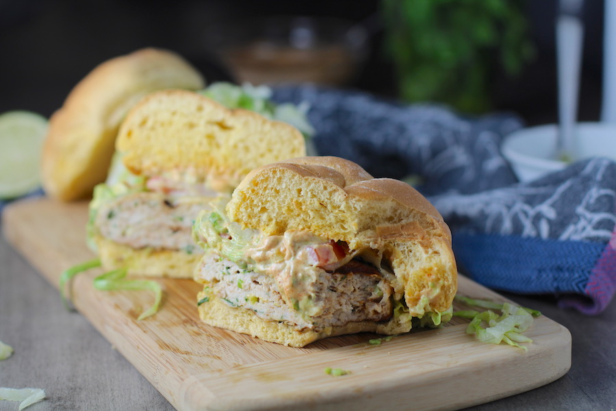Taco Chicken Burger cut in half with a bite taken out on wood plate. The burger has smokey taco seasonings, cilantro, and scallions. Then on top are melty cheese, guacamole, lettuce, tomato, and chipotle mayonnaise!