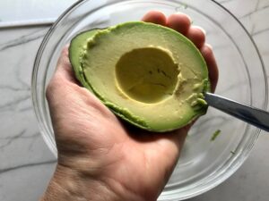 Hand holding half avocado and spoon scooping it out for a Taco Chicken Burger recipe. The burger has smokey taco seasonings, cilantro, and scallions. Then on top are melty cheese, guacamole, lettuce, tomato, and chipotle mayonnaise!