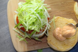 Taco Chicken Burger on wood platter with top bun off to side. The burger has smokey taco seasonings, cilantro, and scallions. Then on top are melty cheese, guacamole, lettuce, tomato. On the top bun is chipotle mayonnaise!