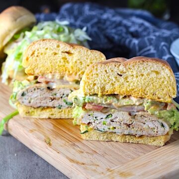 Chicken or Turkey Taco Burger cut in half on wood plate. The burger has smokey taco seasonings, cilantro, and scallions. Then on top are melty cheese, guacamole, lettuce, tomato, and chipotle mayonnaise!