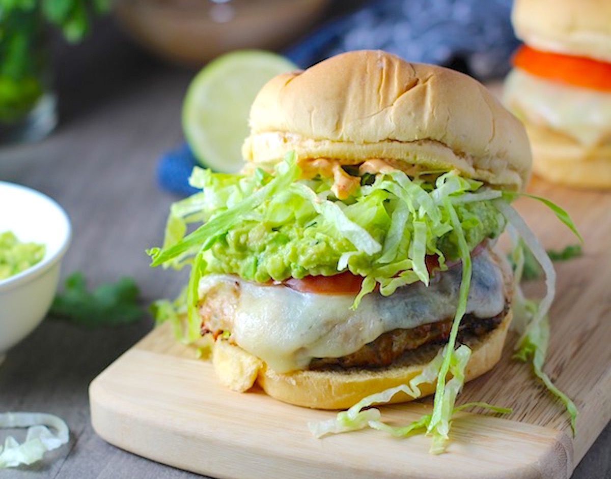 Chicken or Turkey Taco Burgers on wood platter with smokey taco seasonings, cilantro, and scallions. Then on top are melty cheese, guacamole, lettuce, tomato, and chipotle mayonnaise!