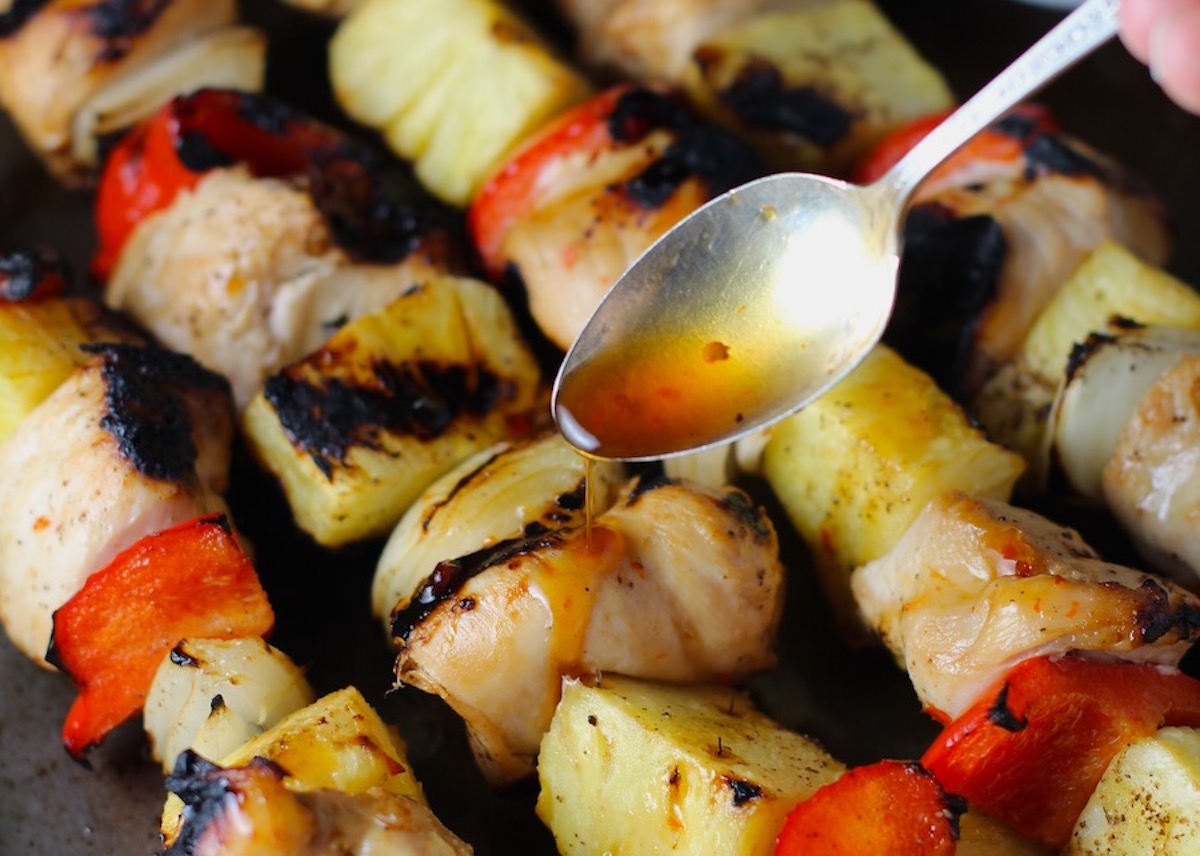 Spoon drizzling Sweet and Sour sauce over Chinese Chicken and Pineapple Kabobs with onion, and red pepper on sheet pan.