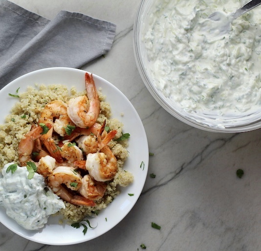 Tzatziki sauce in a large mixing bowl next to a plate of shrimp over quinoa with tzatziki. This is a delicious and easy Tzatziki Recipe with cool and creamy yogurt, fresh cucumber, lime zest, garlic, and scallion.