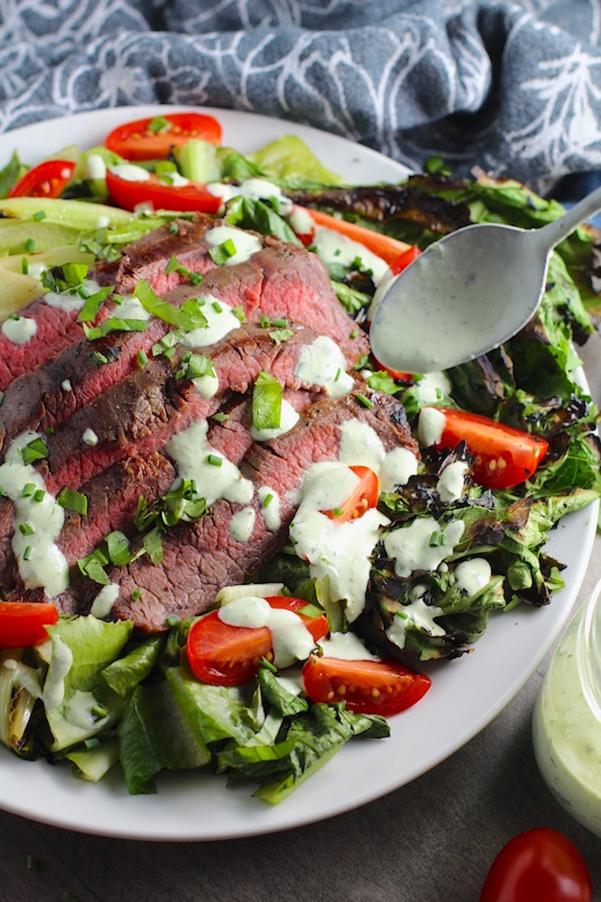 Spoon drizzling dressing over sliced grilled Flank Steak Salad Recipe on plate with grilled romaine, halved grape tomatoes, and creamy basil yogurt dressing.